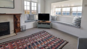A room with a fireplace, a television on a wooden stand, a patterned rug, and a window seat with pillows. - 4 East Cliff (Lyme Regis)