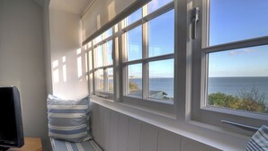 A room with a large bay window overlooking the sea, a television on a wooden table, and a striped bench. - 4 East Cliff (Lyme Regis)