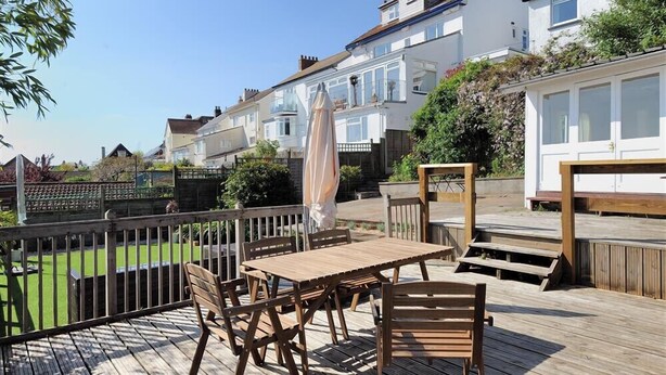 Une terrasse en bois avec une table et des chaises, entourée d’une clôture blanche et avec des maisons en arrière-plan.