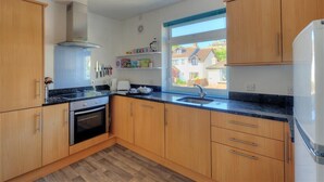 Une cuisine moderne avec des meubles en bois, un four encastré, une hotte en acier inoxydable, un plan de travail noir et une fenêtre donnant sur les maisons à l’extérieur.