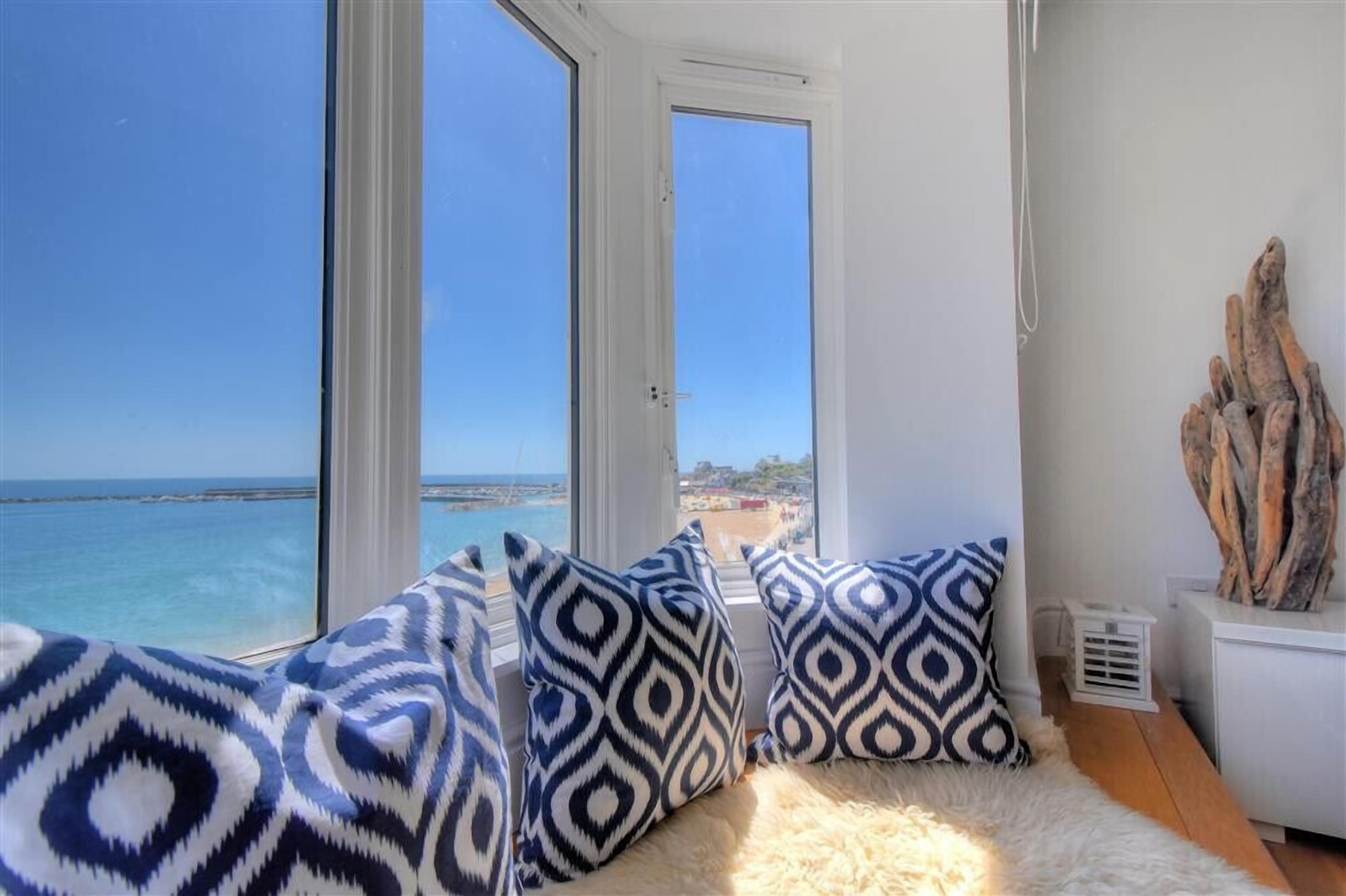 Una habitación con un amplio ventanal con vista a la playa, un tapete blanco y mullido, y cojines estampados.