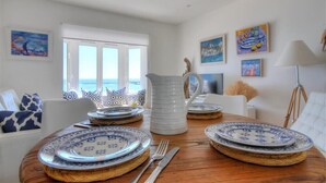 A dining area with a wooden table set for four, white walls, and a view of the sea through large windows. - Seahorse (Lyme Regis)