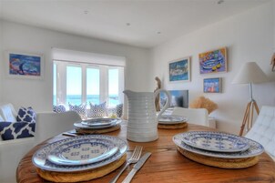 Comedor con mesa de madera dispuesta para cuatro personas, paredes blancas y vista al mar a través de amplios ventanales.