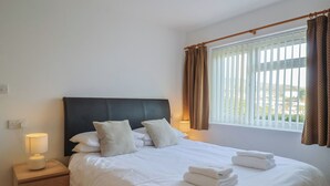 A neatly made bed with white linens and pillows, a bedside lamp, and a window with curtains. - Greystones Flat (Lyme Regis)