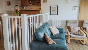 A living room with a teal leather sofa, wooden chairs, a white railing, and a kitchen in the background. - Greystones Flat (Lyme Regis)
