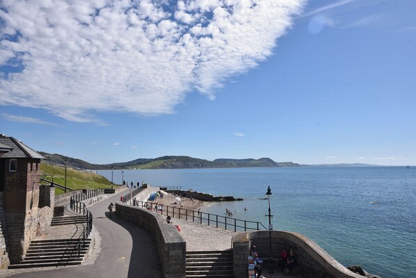 Une promenade en bord de mer avec des escaliers menant à une plage, un mur de pierre et une vue dégagée sur la mer et les collines lointaines.