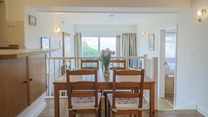 A dining area with a wooden table and chairs, a vase with flowers, and a view of the outdoors through large windows. - Greystones Flat (Lyme Regis)