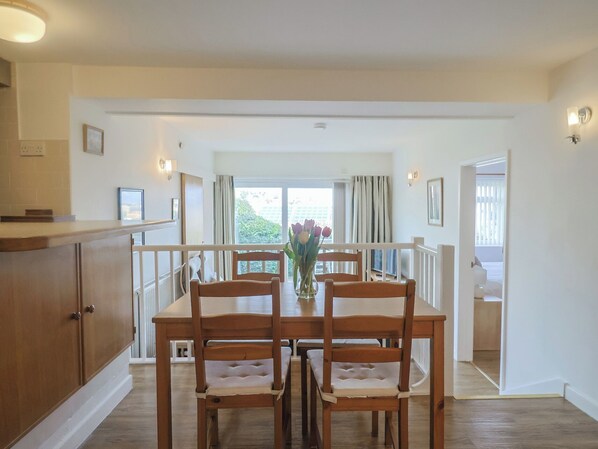 A dining area with a wooden table and chairs, a vase with flowers, and a view of the outdoors through large windows.