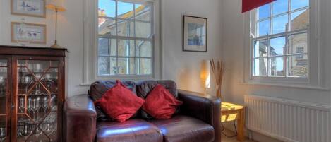 A cozy living room with a leather sofa, a wooden cabinet with glass doors, and two windows with red valances.
