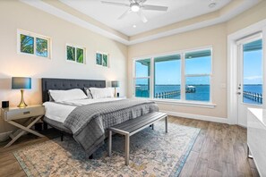 Une chambre à coucher comprenant un lit, des tables de chevet, un banc, une grande fenêtre donnant sur la mer et des œuvres d’art accrochées au mur.