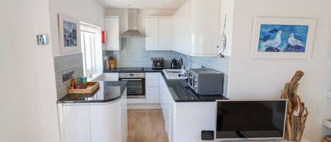 A modern kitchen with white cabinets, a built-in oven, a microwave, and a toaster. There is a wooden dining table with a TV and a remote control.