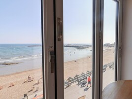 En utsikt över en strand från ett rum med ett stort fönster, med havet, stranden och en avlägsen pir i bild.