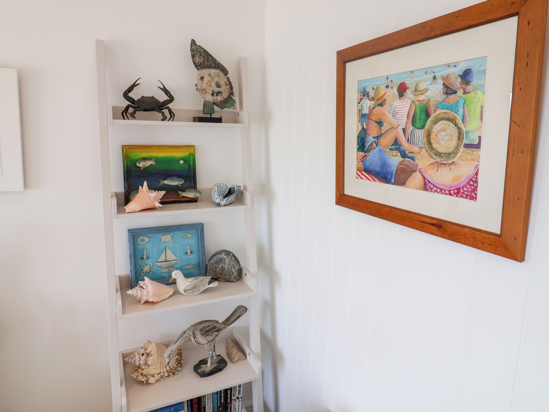 A room with a white wall and a wooden-framed painting of beachgoers. Shelves contain various decorative items including seashells, a crab, and a fish sculpture.
