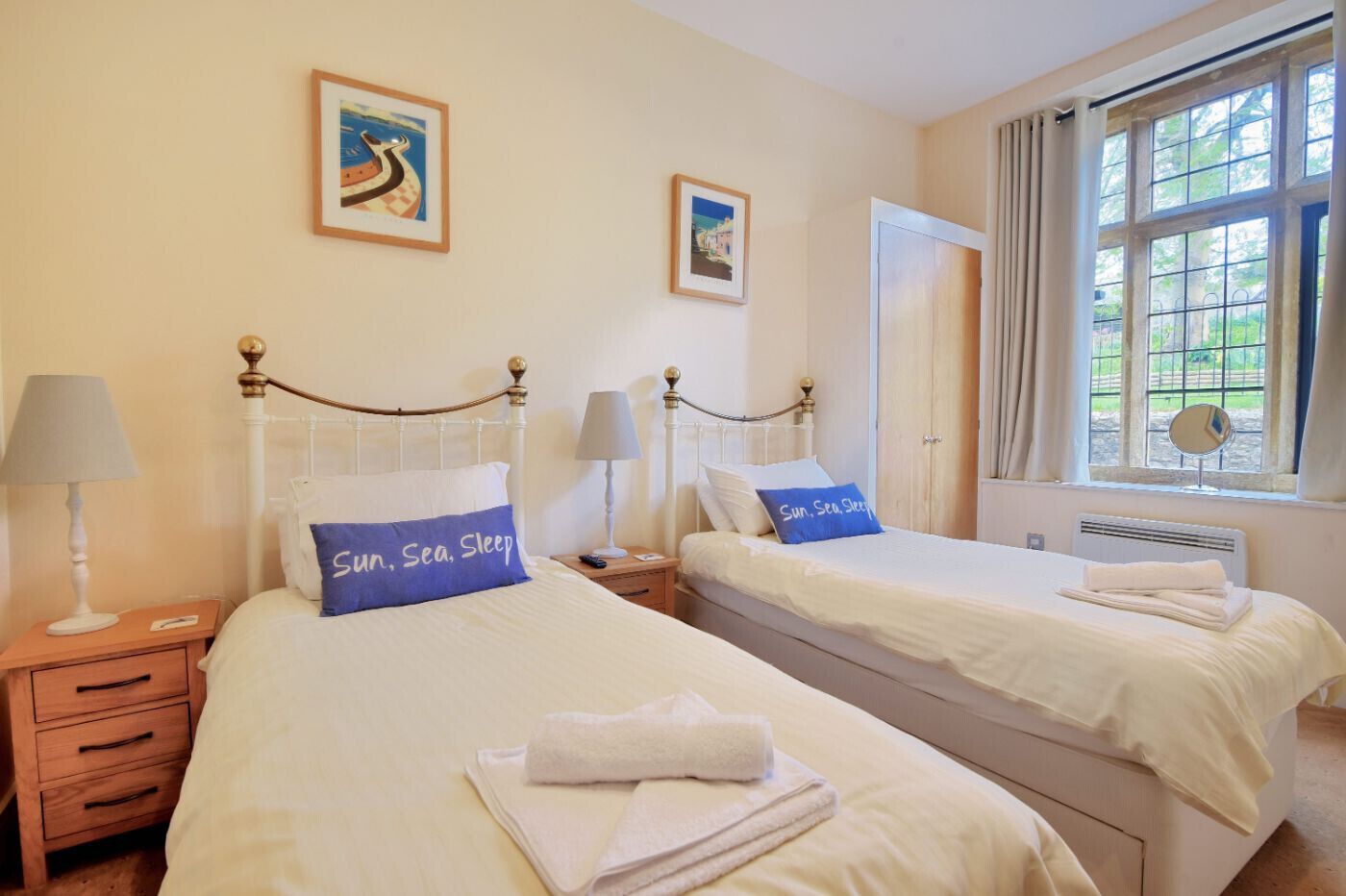 A hotel room with two beds, each with a pillow labeled "Sun, Sea, Sleep." There are bedside tables with lamps, a window with curtains, and a wall with two framed pictures.