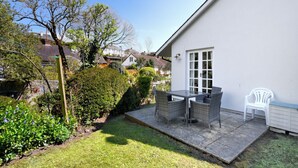 Une terrasse avec une table et des chaises, un mur blanc et un jardin agrémenté d’arbustes et d’arbres.