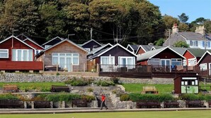 A row of wooden cabins with stone walls, a grassy area with benches, and a person walking. - 8 Bowling Green Chalets (Lyme Regis)