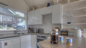 A kitchen with white cabinets, a stove, a sink, and a window with blinds. - 8 Bowling Green Chalets (Lyme Regis)