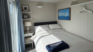 A bedroom with a bed, a nightstand, a lamp, a picture frame, a towel, and a hanging clothes rack. - 8 Bowling Green Chalets (Lyme Regis)