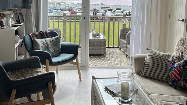 A living room with a TV, two chairs, a sofa, and a balcony with a view of parked cars and a grassy area. - 8 Bowling Green Chalets (Lyme Regis)
