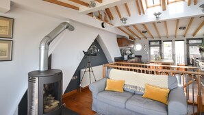A modern loft with exposed wooden beams, a fireplace, a cozy seating area, and a kitchen with large windows. - The Lookout (Lyme Regis)