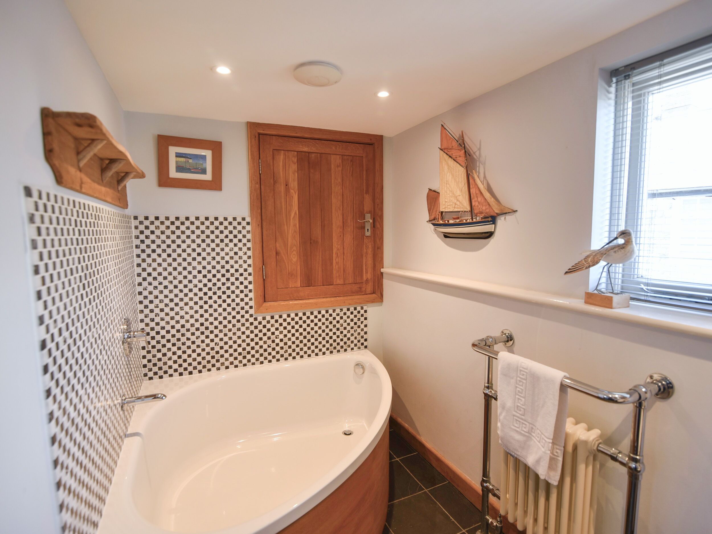 A bathroom with a freestanding bathtub, a wooden cabinet, a ship decoration, and a window with blinds.