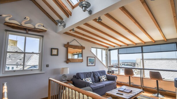 A spacious living room with a high ceiling, wooden beams, a large window with a view of the sea, a sofa, a coffee table, and a small table with a lamp. - The Lookout (Lyme Regis)