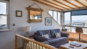 A cozy living room with a wooden ceiling, a large window with a view of the city, a grey sofa with patterned cushions, and a wooden coffee table. - The Lookout (Lyme Regis)