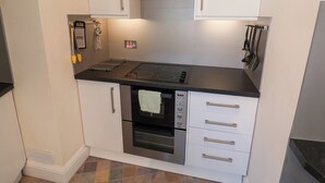 Une cuisine compacte avec des meubles blancs, un four encastré, une plaque de cuisson et divers ustensiles accrochés au mur.