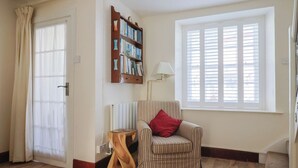 A living room with a striped armchair, a wooden side table, a bookshelf, a window with blinds, and a door with curtains. - 3 Dolphin Cottages (Lyme Regis)