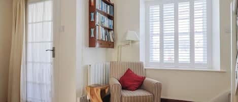 A living room with a striped armchair, a wooden side table, a bookshelf, a window with blinds, and a door with curtains.
