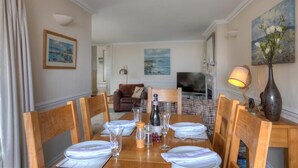 A dining area with a wooden table set for six, a brown sofa, and a TV in the background. - The Portland Suite (Lyme Regis)