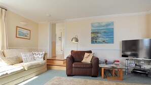 A living room with a beige sofa, a brown leather chair, a glass coffee table, a flat-screen TV, and a painting of a beach scene. - The Portland Suite (Lyme Regis)