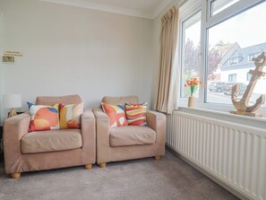 Une pièce avec deux fauteuils beiges, des coussins colorés, un radiateur et une fenêtre donnant sur une maison et des voitures garées.