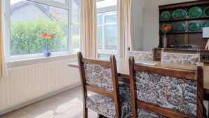 Uma área de refeições com mesa e cadeiras, uma janela com cortinas e vista para o verde lá fora.