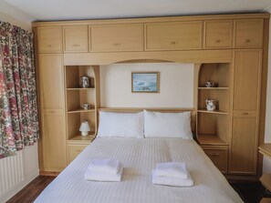 Quarto de hotel com cama grande, mesa de cabeceira, luminária e guarda-roupa com várias prateleiras.