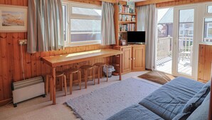 A cozy cabin with a wooden interior, a dining area with a table and chairs, a bookshelf, a television, and a large window overlooking a deck. - Granny's Teeth (Lyme Regis)