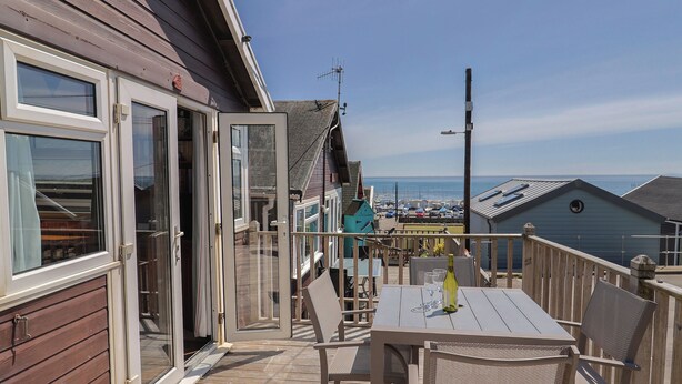 A wooden deck with a table and chairs, a sliding glass door, and a view of the sea and beach in the background. - Granny's Teeth (Lyme Regis)