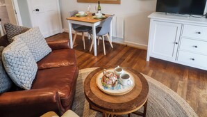 A living room with a brown leather sofa, a round wooden coffee table with a teapot and cups, a dining table with chairs, a white cabinet with a TV, and a framed artwork on the wall. - The Apartment at Queen Anne's Lodge (Lyme Regis)