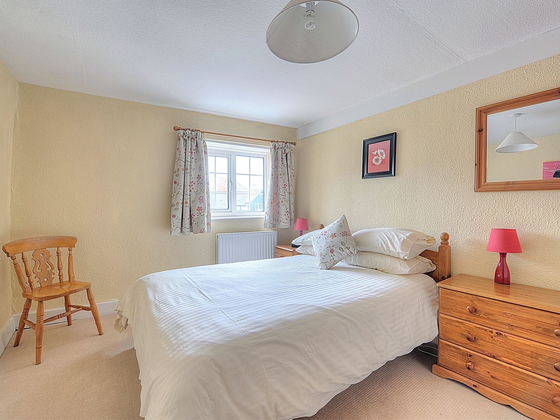 A bedroom with a bed, a wooden chair, a dresser, a window with curtains, a radiator, and a framed picture on the wall.