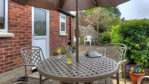 Une terrasse avec une table ronde et des chaises, un grand parasol et un mur en briques avec une fenêtre.
