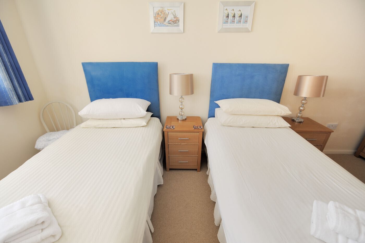 A hotel room with two single beds, each with a blue headboard, white bedding, and a bedside table with a lamp. There are two framed pictures on the wall above the beds.