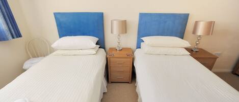 A hotel room with two single beds, each with a blue headboard, white bedding, and a bedside table with a lamp. There are two framed pictures on the wall above the beds.