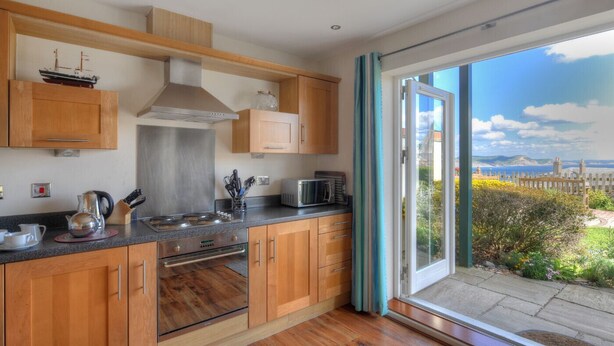 A modern kitchen with wooden cabinets, stainless steel appliances, and a view of a coastal landscape through a large sliding glass door. - 8 Buckfields (Lyme Regis)