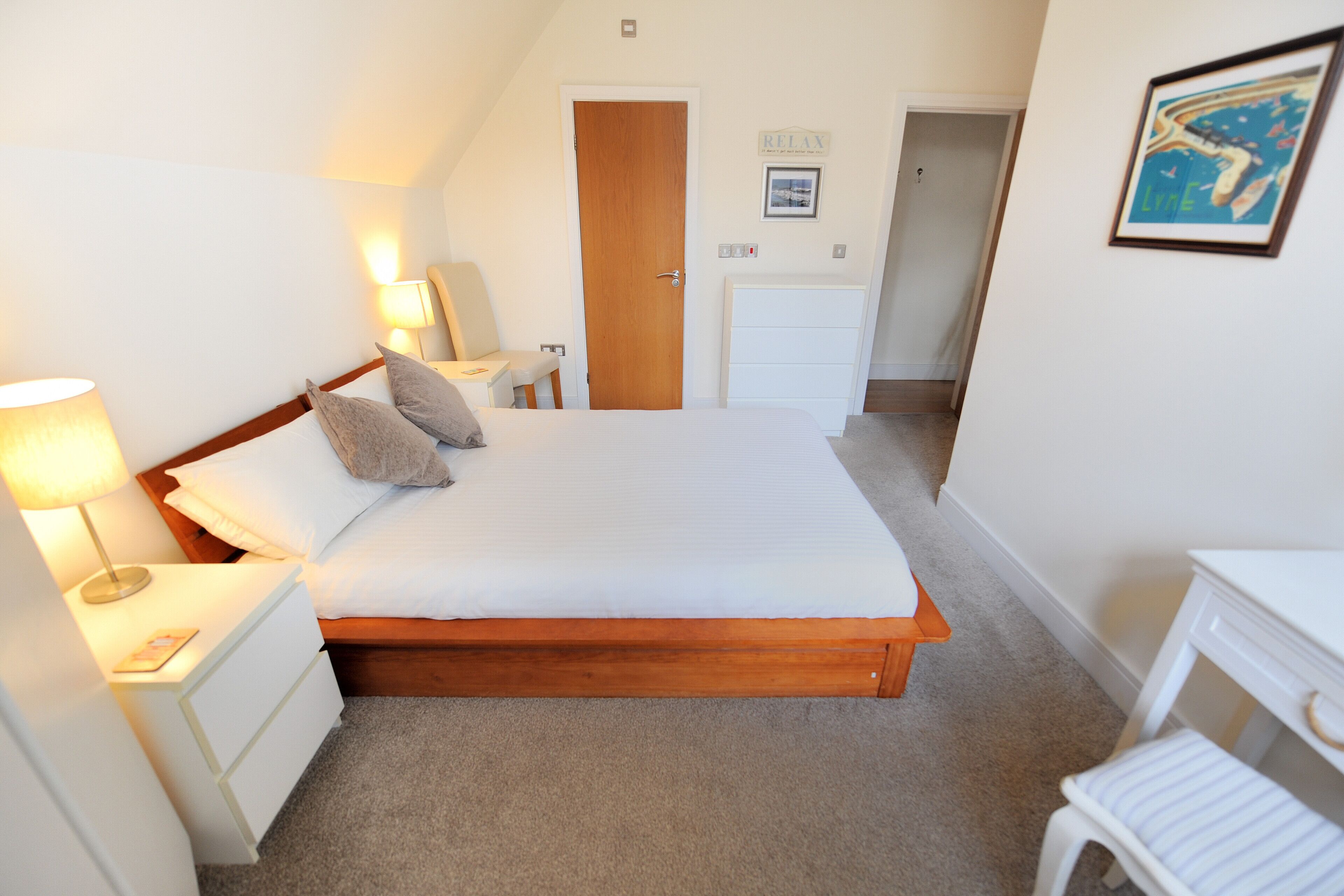 A bedroom with a wooden bed, white bedside tables, a nightstand, a lamp, a small chair, and a desk with a chair.