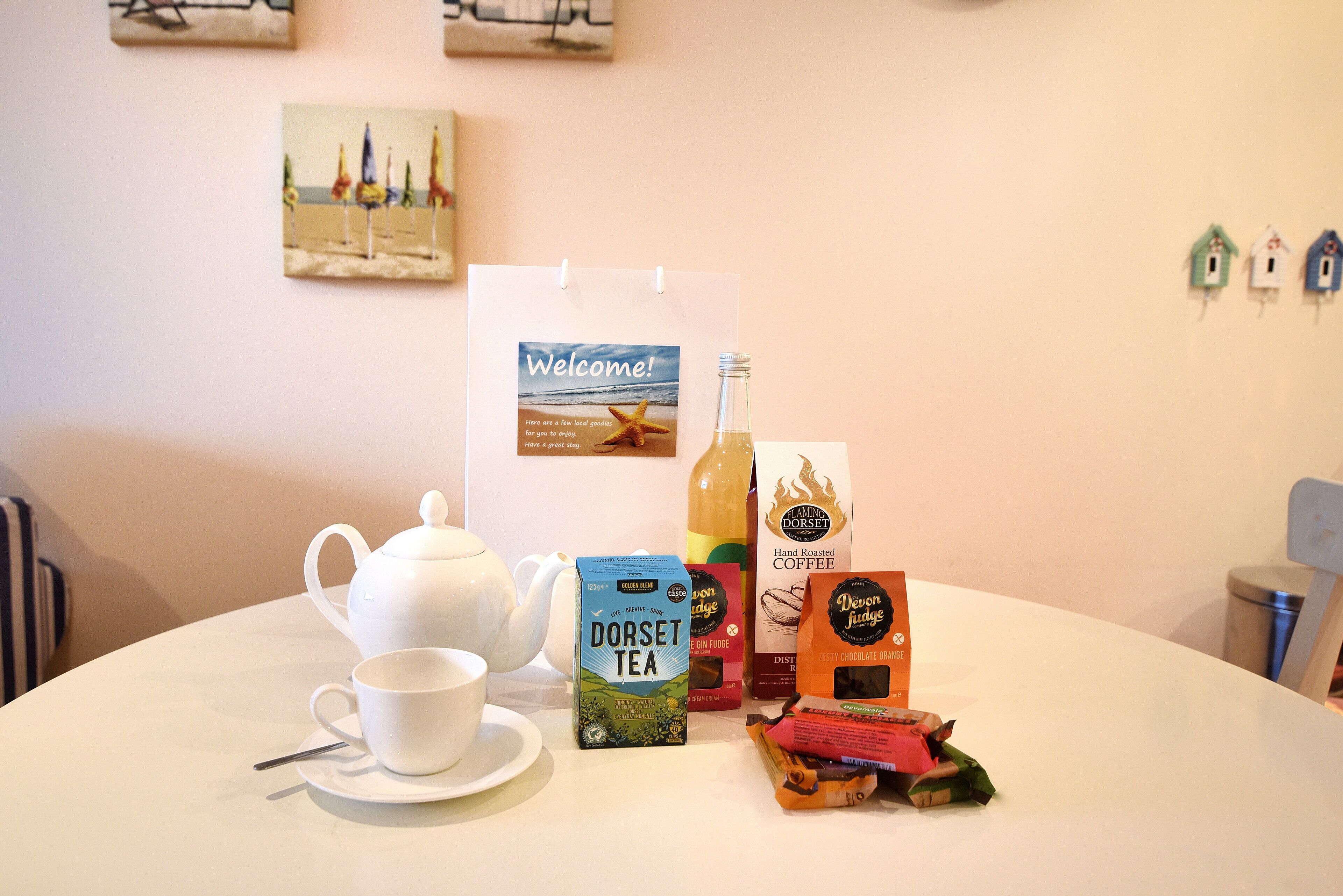 A table with a teapot, cup, and various food and drink items, including tea and coffee, in a room with a simple decor and a welcome sign on the wall.