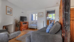 A living room with a grey sofa, a television, a wooden cabinet, and a window with curtains. - Ivy Cottage (Lyme Regis)
