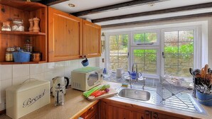 A kitchen with wooden cabinets, a bread box, a kettle, and a window with a view of greenery. - Ivy Cottage (Lyme Regis)
