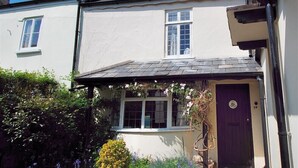 A two-story house with a white exterior, a dark roof, and a purple door. There is a small garden with various plants and flowers in front of the house. - Ivy Cottage (Lyme Regis)