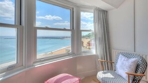 A room with a view of the sea and a beach, featuring a window with curtains, a patterned chair, and a pink cushion. - Ross House (Lyme Regis)