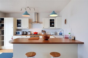 A modern kitchen with white cabinets, a wooden island, two bar stools, a microwave, a kettle, and a red pot. - Ross House (Lyme Regis)